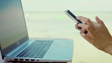 Sahilde şapkalı bir kadın dizüstü bilgisayar kullanıyor, akıllı telefon kullanıyor, internette sohbet ediyor, internette geziniyor.