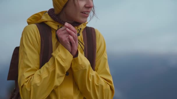 Крупным планом портрет замерзшей украинки в ярко-желтом плаще и шапочке, с рюкзаком стоит на открытом воздухе в холодный облачный осенний день. Девушка пытается согреть свои замороженные руки собственным дыханием, люди — стоковое видео