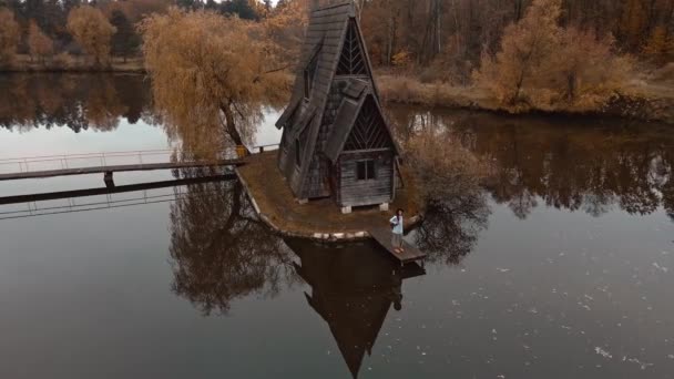 空中俯瞰女旅行家站在一座古老的木制小屋旁边，湖边林立着红叶的树木，秋天的自然景观。乌克兰的自然美景和旅游景点. — 图库视频影像