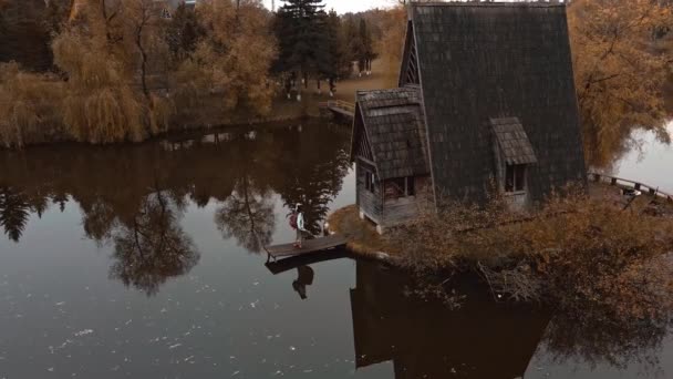 空中俯瞰女旅行家站在一座古老的木制小屋旁边，湖边林立着红叶的树木，秋天的自然景观。乌克兰的自然美景和旅游景点. — 图库视频影像