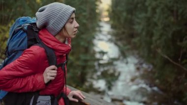 Ağır çekimde genç bir kadın yürüyüşçü ahşap köprüde duruyor ve dağ nehrinin üzerinde asılı duruyor. Çayırları, Ukrayna 'daki dağları olan muhteşem bir manzara..