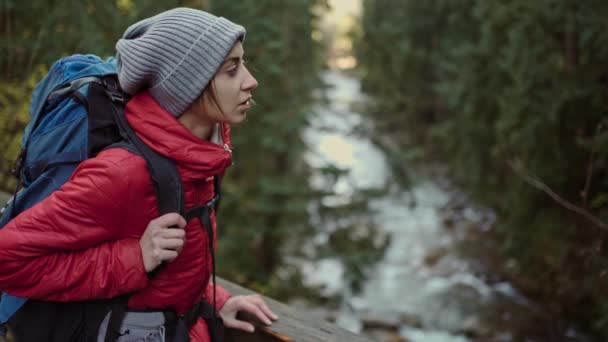 Retrato en cámara lenta mujer joven excursionista de pie en el puente de madera colgado sobre el río de montaña. paisaje increíble con abetos, montañas en Ucrania. — Vídeos de Stock