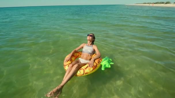 Счастливая улыбающаяся молодая женщина в солнечных очках, плавающая на надувном ананасе, плавающем кольце в морской воде и смеющаяся. Камера уменьшается. путешествия и летние каникулы концепции. Украина, Одесская область. — стоковое видео