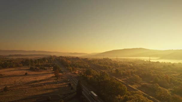 Visão aérea cinematográfica fro drone na estrada com veículos de carga e ferrovias no nascer do sol ensolarado no vale da montanha em Cárpatos, oeste da Ucrânia. Transporte e entrega de carga. — Vídeo de Stock