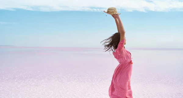 En ansiktsløs frihetskvinne i rosa kjole med hatt over hodet, stående på en øde strand i rosa innsjø. – stockfoto