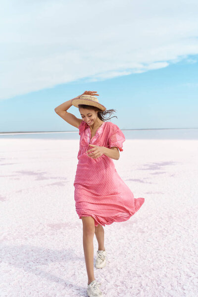 Fabulous woman walking by beach of pink lake. Girl in hat and pink dress blowing Royalty Free Stock Photos