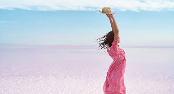 Faceless freedom woman in pink dress holding hat over head, standing on desert beach of pink lake. Stock Photo