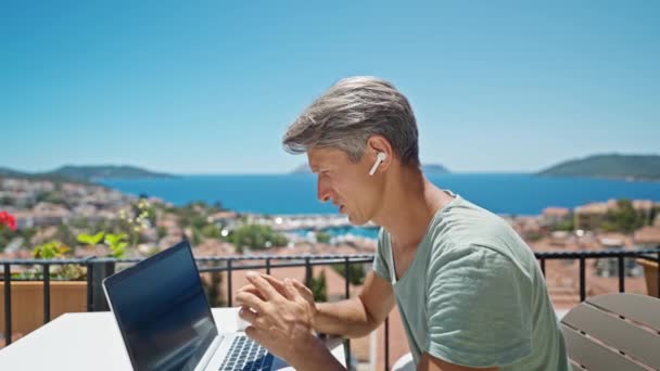 ドルリーは美しい青い海の景色によって屋外ノートパソコンで作業しているイヤホンの映像男をズームアウトし オンラインビデオ通話で話しています コンセプトリモートワークと旅行 — ストック動画