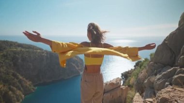 Ağır çekimde özgürlük, kollarını kaldıran bir kadın Kelebek Vadisi 'nin güzel manzarasının keyfini çıkarıyor. Sarı tişörtlü çekici kız denizin üzerinde uçurumun tepesinde duruyor, saçları rüzgarla savruluyor.