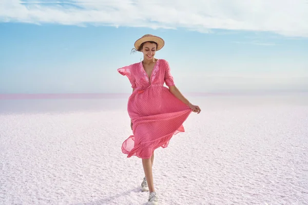 Elegant girl in flowing pink dress enjoying walk by beautiful landscape of salt flats on pink lake with blue cloudy sky, looks like white desert.