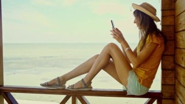 Plajda şapkalı bir kadın, akıllı telefon kullanıyor, internette sohbet ediyor, internette geziniyor. Teknoloji ve seyahat, tatilde online iletişim..