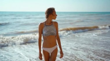 Bikinili genç fitness kızının yavaş çekimde çekilmiş portresi deniz sahilinde dalgalarla yürüyor. İyimserlik, iyimserlik, tasasız, sağlıklı vücut pozitif ve refah mutluluk kavramı.