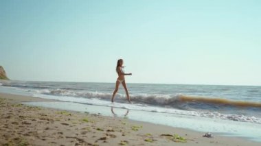 Mutlu, neşeli bir kadın, deniz kıyısında atlıyor ve koşuyor. Sağlığını ve özgürlüğünü hissediyor. Bikinili neşeli kız dışarıda eğleniyor..