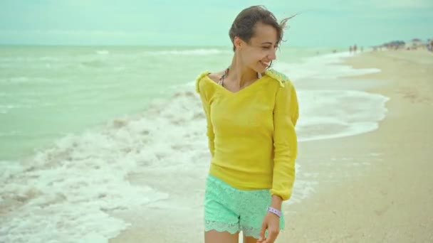 本物のダイナミックなポートレート幸せな表情の女性が吹いて髪を身に着けています黄色のシャツビーチで楽しいと楽しいカメラの周りを実行している — ストック動画