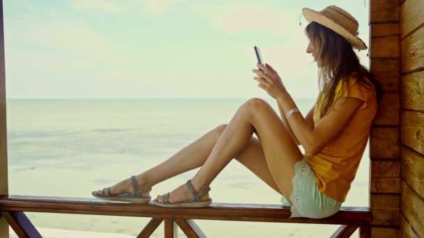 边看沙滩上戴帽子的女人 用智能手机 网上聊天 上网浏览 技术和旅行 度假时在线交流 — 图库视频影像