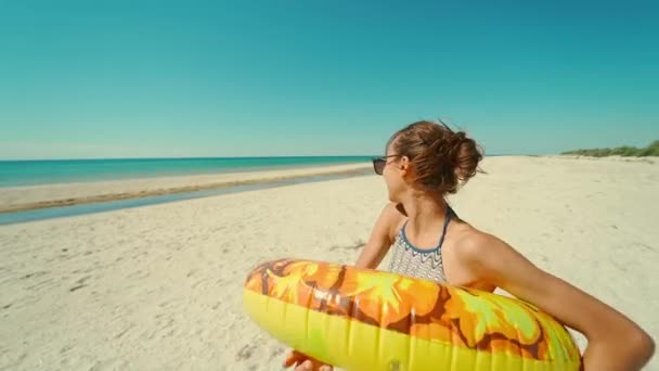 太陽の光が差し込む海のビーチでカメラを見る笑顔の女の子面白い顔をした黄色のインフレータブルリング 夏に白砂のビーチで楽しいビキニの幸せな若い女性 女性は顔を近づけて — ストック動画