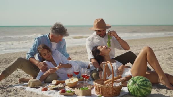 在明媚的日子里 两对年轻貌美的情侣在沙滩上举行浪漫的野餐 人们一起庆祝 用食物喂对方 — 图库视频影像