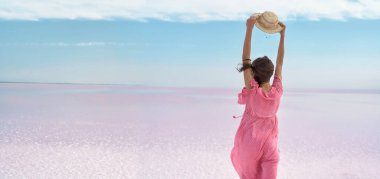 Pembe elbiseli zarif bir kadının mavi gökyüzüne karşı şapkasını tutarken, pembe göl tuzu düzlüklerinin çöl ufkunda dikilirken görüntüsü panoramik.