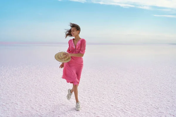 Elegant woman walking by windy serenity landscape of salt flats on pink lake. Motion image of girl in pink flowing dress with blowing hair making step
