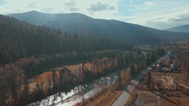 Aerial countryside ukrainian village in Carpathian mountains. Autumn rural landscape. Drone flies over houses, road, river and pine tree forest, mountain range in background. Wild nature scene.