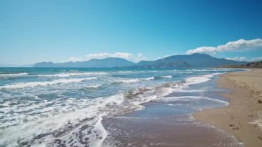 slow motion camera movement along waving sea with waves at sunny summer windy day at beautiful sandy empty beach Iztuzu in Turkey. beautiful mountain range on background