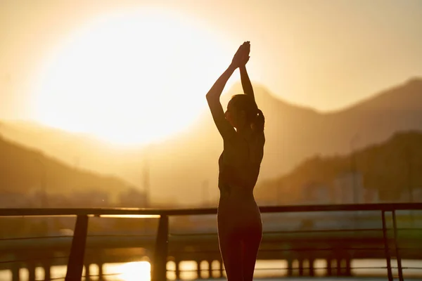 Silhouette woman yoga on sunset with hands up. Practicing meditation and yoga on the bridge against sea water and mountain range