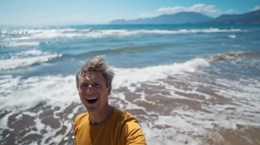 happy middle aged greyhead man takes selfie at sea coast with waves enjoying sunny day on beautiful beach Iztuzu in Turkey. inspired enjoyed man makes video call and talks about his place