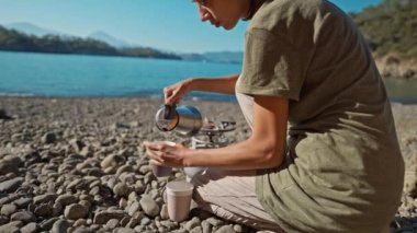 Kadın yürüyüşçü turist kahve makinesinden bardaklara kahve doldurur Akdeniz kıyılarında çakıl taşlı sahilde hindi ile. Doğada gün doğumu ve sabah içeceği. Çadır yoluyla sinematik görünüm