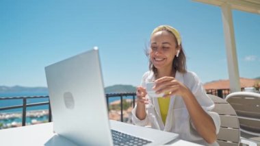 Çekici melez kadın, Türk tatil beldesindeki deniz manzaralı terasta oturup sabah kahvesini yudumlarken, bilgisayar kullanarak ünlü veya uzak çalışma, serbest çalışma kavramına bilgisayar kullanıyor. Yavaş çekim 4k