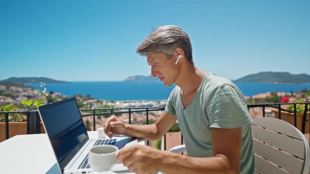 ドルリーは美しい青い海の景色によって屋外ノートパソコンで作業しているイヤホンの映像男をズームアウトし オンラインビデオ通話で話しています コンセプトリモートワークと旅行 — ストック動画