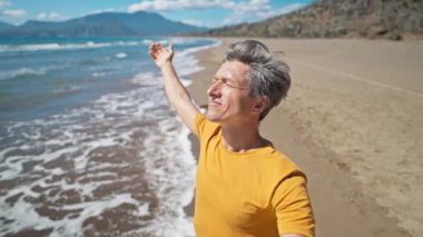 close up selfie portrait of happy middle aged greyhead man at sea coast enjoys sunny day on beautiful sandy beach Iztuzu in Turkey. inspired enjoyed man makes video call and talks about his place