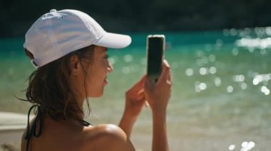 Bikinili neşeli, bronz tenli kadın deniz kıyısında mavi su arka planında oturuyor ve fotoğraf ya da video çekmek için cep telefonunda kamerasını kullanıyor. Mavi Göl beldesi, Oludeniz, Türkiye.