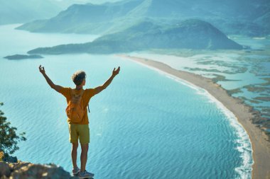 Tepede duran kolları havada bir yürüyüşçü. Başarılı bir adam dağlarla ve deniz kıyılarıyla güzel manzaranın tadını çıkarıyor.