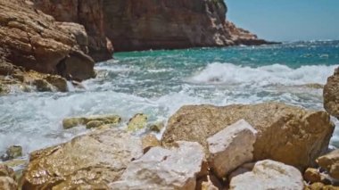 Akdeniz kıyısındaki kayalık çakıl taşı plajına ağır çekim deniz dalgaları çarpıyor ve köpürüyor. Altın kayalar derin mavi okyanus suyuyla buluşuyor. Türkiye 'de Yaz Turizmi