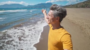 happy middle aged greyhead man stands at sandy sea coast with raised hands. tourist enjoys sunny day and fresh air on beautiful empty beach Iztuzu in Turkey. travel and vacation concept