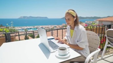 Young elegant woman with wireless earphones looking at laptop with smile while sitting at balcony with seaview on resort. girl freelancer works outdoors during travel