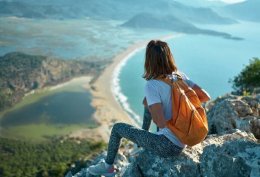 Back view tourist woman sitting on rock cliff and admiring amazing scenic nature view over sea coast with mountains range