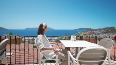 camera movement around young mixed race smiling woman sitting on open terrace with beautiful sea view in hotel at turkey resort. go everywhere