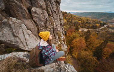 Woman tourist in yellow hat with backpack sitting on cliff edge with autumn forest and valley view. Adventure vacations tour outdoor to national park in Ukraine, Tustan.