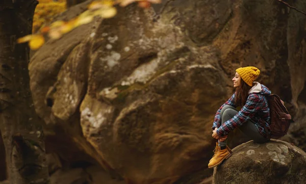 Side portrait of woman relaxing in the nature. Millennial girl in checkered jacket and beanie hat with backpack sits on stone and enjoying silence and good nature vibes alone