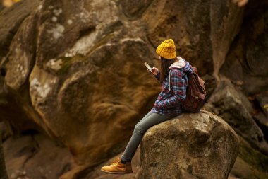 Young woman hiker in checkered jacket and beanie with backpack sits on big boulder over forest and using smartphone, chatting