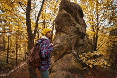 Young hipster woman in checkered jacket and yellow beanie hat with backpack enjoying walk in autumn golden forest with mossy boulders in Carpathian, Ukraine.