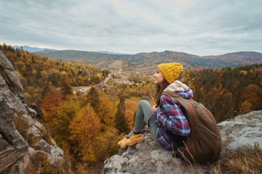Traveler millennial woman in checkered jacket and yellow beanie hat with backpack sitting on cliff edge over mountains with fall nature view