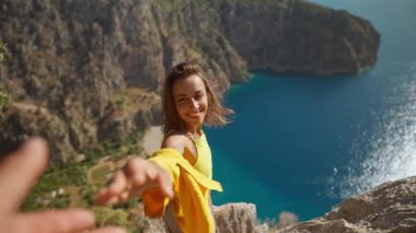 slow motion first person view happy smiling cheerful young woman tourist in yellow shirt stands on cliff edge over sea. girl enjoys summer travel in Turkey and take hand a man