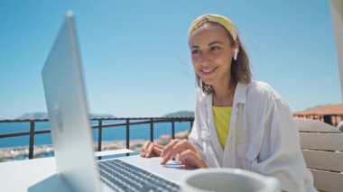 Young elegant woman with wireless earphones looking at laptop with smile while sitting at balcony with seaview on resort. girl freelancer works outdoors during travel