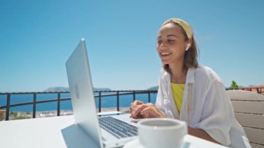 Happy woman in earphones communicating by conference call speak sitting outdoors on summer terrace with beautiful sea view, video chat job interview or distance language course clas online teacher