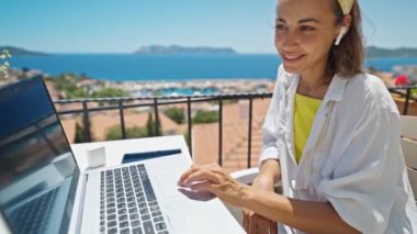 4k slow motion of beautiful young freelancer woman working on laptop outdoors on terrace at hotel resort, in wireless earphones enjoying beautiful sea view and sunny morning