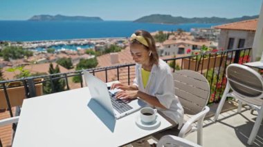 4k slow motion of beautiful young freelancer woman working on laptop outdoors on terrace at hotel resort, in wireless earphones enjoying beautiful sea view and sunny morning