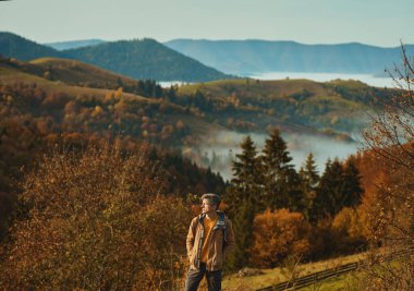 Man hipster hiker looking around, enjoying the view above the clouds on meadow of mountain ridge against mountains and woods.Carpathian mountains, Ukraine