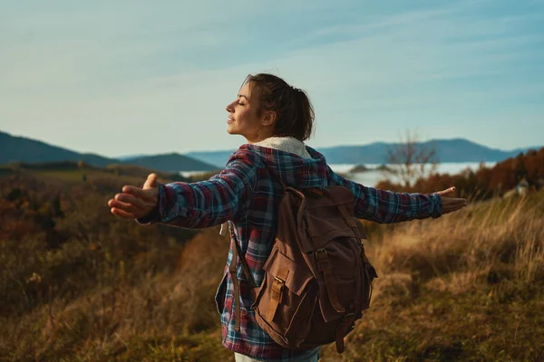 Rear view girl hiker wearing checkered jacket and backpack, standing with open arms, enjoying beautiful view over mountain